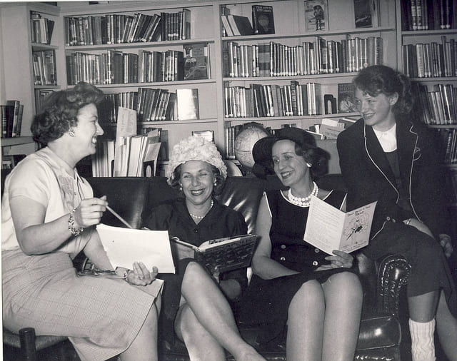 Four women in a library, two reading books, in a history backup context.