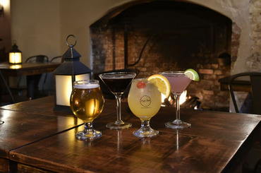 Four craft cocktails and beer on a table at The 1747 Pub with a fireplace