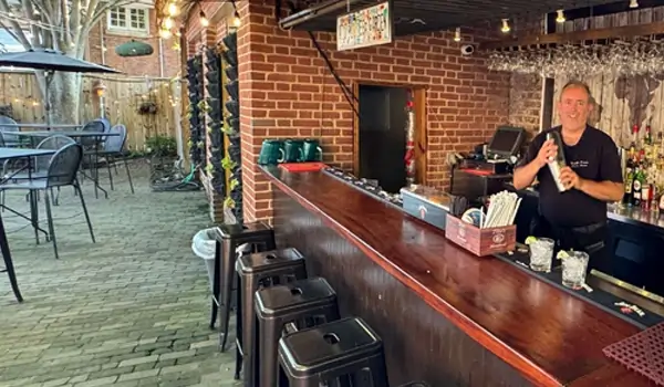 Bartender at The 1747 Pub's outdoor bar with brick wall and seating.